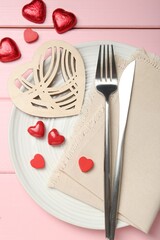 Romantic place setting for Valentine's day. Cutlery, napkin, plate and decorative hearts on pink wooden table, flat lay