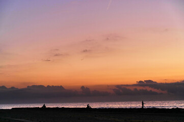 As the sun sets, silhouettes of people stroll along the tranquil shore, with soft waves lapping against the beach and colors painting the sky.
