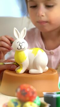 Mother and daughter paint a ceramic bunny together for Easter. The girl, wearing bunny ears, carefully applies yellow paint while her mother assists. The table is filled with colorful paints