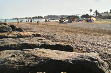 Families and friends gather on a sunny beach, basking in the warmth while children play in the sand and waves. Rocky formations add a natural beauty to the scenic coastline.