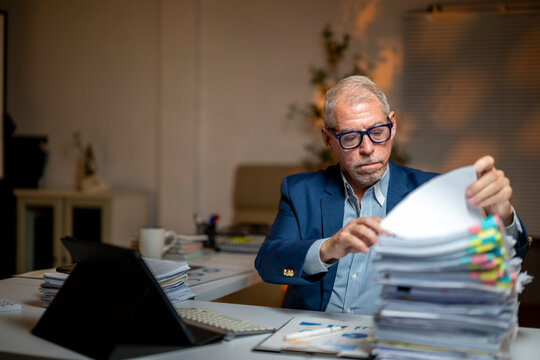 A man in a suit is looking at a stack of papers on his desk