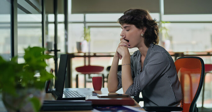 Young adult gen z woman lady sitting use laptop do over work time look screen feel bad lazy tired lack dull job office place busy sleepy teen female bored yawning on desk table need break rest