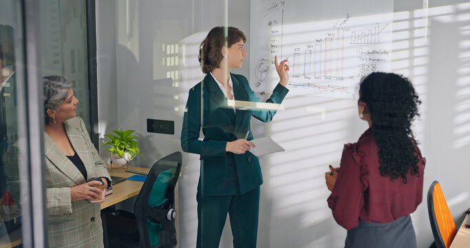 Young adult boss female leader talk share stock idea show growth chart graph data on white board place old elder older lady teen girl intern staff team expert group listen work plan job office glass