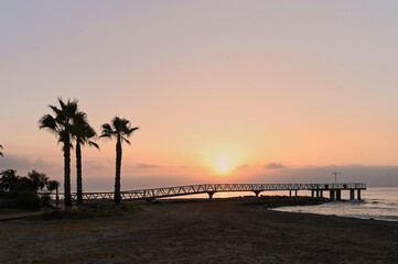 Bright golden rays pierce the horizon, casting a warm glow over the pier extending into the calm...