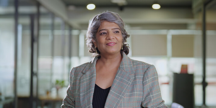 Indian happy old aged boss ceo woman lady do routine spend break time standing in hall room space smiling elder older relax agent worker staff enjoy day office job work place