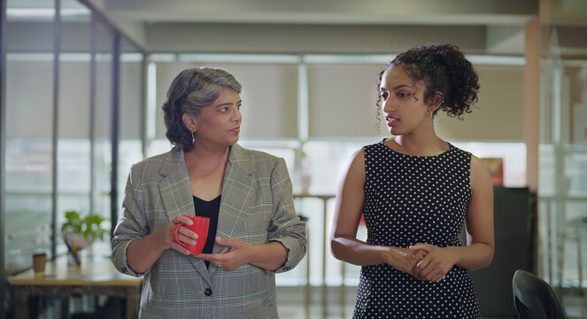 Indian Asian senior boss women lady drink hold coffee cup mug gossip talk indoor work place young adult teen curly hair girl speak share idea listen plan during tea break spend time enjoy job day