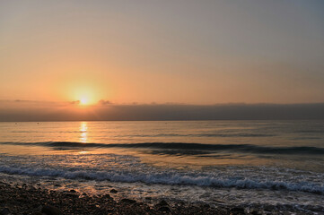 As the sun sets on the horizon, warm golden hues paint the sky while gentle waves kiss the pebbled shore, creating a tranquil evening at the beach.