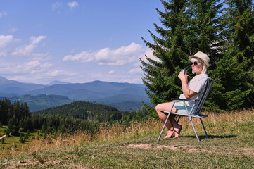 Sitting comfortably in a chair, a woman relaxes against a backdrop of lush green mountains and...