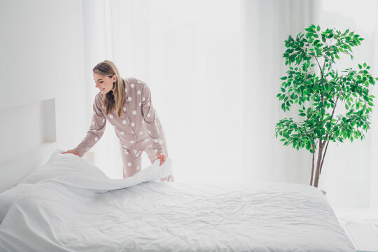 Young woman in cozy pajamas tidying up bed in bright bedroom with natural daylight