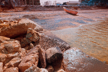 Infrared photography, a cluster of large, weathered rocks with a textured, reddish-brown hue. A lone boat, painted in a vibrant orange, rests on the sand