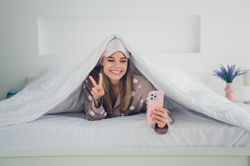 Young woman in pajamas enjoys a peaceful morning in bed, smiling and using her phone under a cozy...