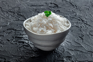 White rice, cooked. A bowl of boiled long grain rice, a simple side dish, with fresh parsley, side view on a black table