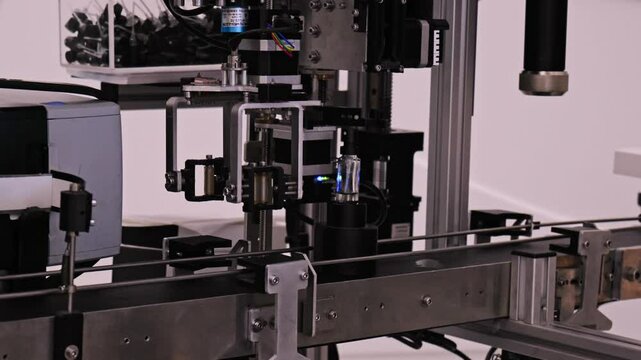 Robot is working to produce product. Robotic arm with suction cup is picking up glass vial from conveyor belt. Human touches glass vial