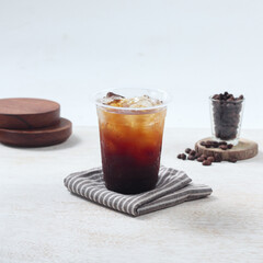 Iced Americano, A clear cup filled with iced coffee sits on a striped napkin, surrounded by coffee beans and wooden coasters in a cozy setting
