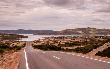 A winding road stretches along a picturesque coast in Teriberka, showcasing a serene lake and distant hills under a cloudy sky, evoking a sense of adventure and tranquility. Travel Russia.