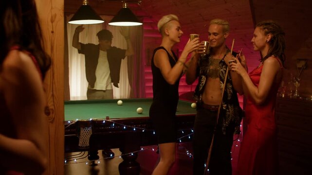 In a stylish room, five young people gather around a billiard table. A man in black and a woman in a red top with white shorts kiss while holding cups. Others cheer amid warm lighting.