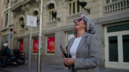 Senior smiling businesswoman in elegant suit walks confidently through the city, holding touchpad. Females determined expression reflects focus on achievement and success in her career and goals
