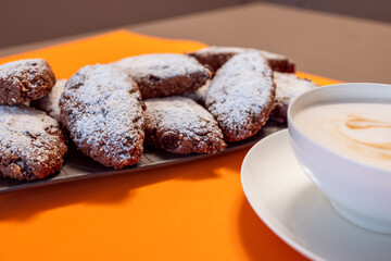 Pan dei Morti cookies with powdered sugar on a dark plate, complemented by frothy cappuccino, set against a vibrant orange background