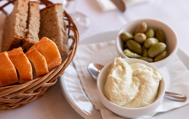 Aioli (allioli) a traditional Mediterranean dip, paired with green olives and an assortment of fresh bread slices presented in a rustic basket on a dining table