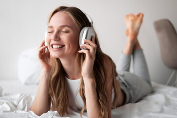 Cheerful young woman listening music with wireless headphones relaxing enjoying online playlist while lying on bed at home.