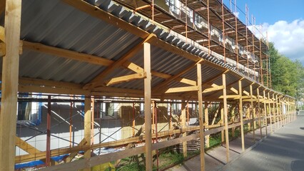 Fototapeta premium Metal scaffolding with wooden decking is installed near the walls of the building for repairs A wooden gallery with a metal roof was installed over the concrete tile sidewalk for pedestrian safety