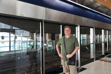 A Middle-Aged Man in Casual Summer Attire Stands Patiently at the Metro Station, Anticipating His...