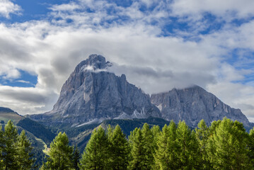 Sassopiatto peak in the Dolomites, Italy, stands over lush meadows and forests, framed by a dramatic sky and steep rocky faces