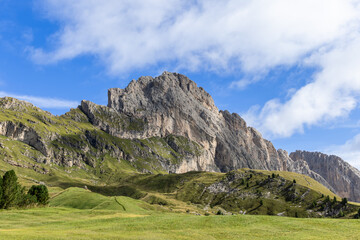 Fototapeta premium The steep rocky slopes of Seceda tower above rolling green meadows, creating a dramatic alpine landscape under a bright blue sky with scattered clouds