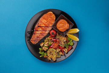 on blue background on a black plate laid out red grilled salmon fillet served with grilled vegetables and additional fish sauce professional cuisine