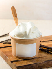 A smooth and creamy vanilla ice cream is served in a paper cup with a wooden spoon on a rustic wooden board alongside whole vanilla beans
