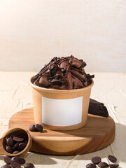 Rich chocolate ice cream is topped with chocolate curls and served in a brown cup on a wooden board beside chocolate chips