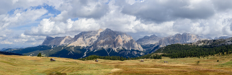 Panoramic view of Pralongia Plateau in the Dolomites, Italy, featuring rolling meadows, scattered alpine huts, and dramatic peaks under a partly cloudy autumn sky