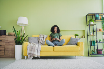 Full length portrait of nice young girl sit sofa use smart phone green interior living room flat...