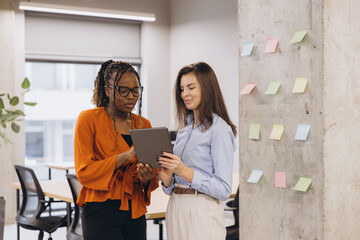Two businesswomen are using a tablet and discussing work next to a concrete pillar with colorful sticky notes in a modern office