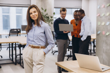 Obraz premium Professional businesswoman smiling confidently, standing foreground, with multicultural team collaborating on laptop project in background