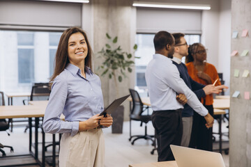 Confident businesswoman holding a clipboard, smiling, with her colleagues discussing in the background in a modern office