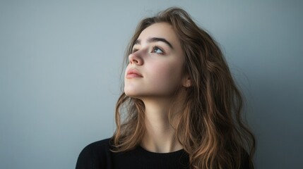 Fototapeta premium Young woman with long wavy brown hair gazing thoughtfully upwards on soft gray background creating a serene contemplative mood in a minimalist setting