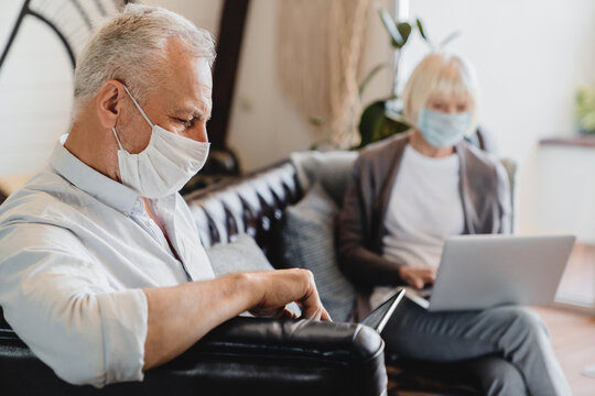 Mature couple sitting with face masks in a bright waiting room or home with laptop and digital tablet. Social distance epidemic pandemic concept. Senior family catch a flu working remotely - Powered by Adobe