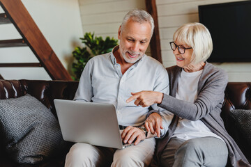 Smiling old Caucasian man and woman spouses use laptop shopping online together form home. Happy mature couple look at computer screen pay bills on internet on gadget. Technology concept.