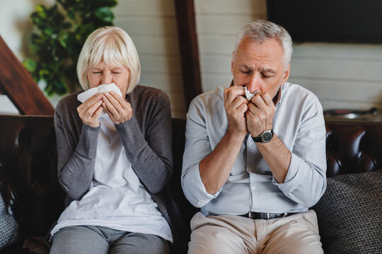 Virus and flu concept. Mature family sitting on sofa and blowing their noses. Sick couple are trying to sneeze in the napkin. They caught a cold and now have to take some medicine to get better.