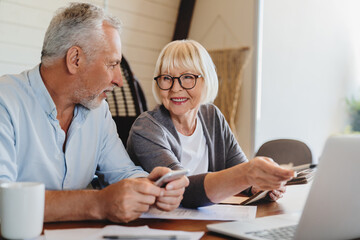 Cheerful elderly grey haired wife and husband sit on couch using computer on-line application...