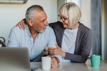 Smiling mature 70s couple sit relax at table at home kitchen have fun shopping online on computer...