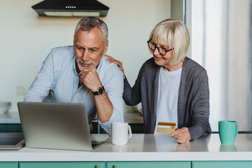 Happy elder retired couple buying goods or booking hotel online, using computer and credit card at...
