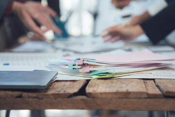 Business Meeting Table Close-up: A selective focus shot of a meeting table, showcasing stacks of papers and a laptop, with blurred figures of colleagues discussing in the background.