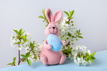 Knitted toy pink bunny with blue Easter egg and with cherry blossoms on blue table and gray...