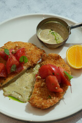 A top-down view of breaded cutlet, topped with a tomato salad and served with a side of sauce