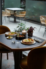 A restaurant table with a pizza, a glass of white wine, and two plates with other food on it. The table is set for two person dinner