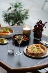 A restaurant table with a pizza, a glass of white wine, and two plates with other food on it. The table is set for two person dinner