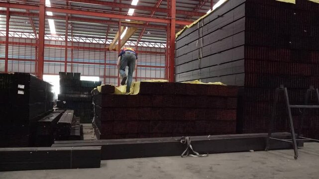 Industrial Wood Material Handling: A worker carefully navigates a stack of lumber within a vast industrial warehouse, surrounded by raw materials and manufacturing equipment, ready for production.