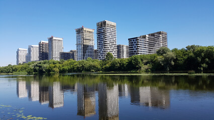 Modern high-rise buildings under construction reflecting in a calm lake with green trees on the shore